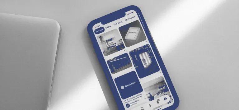 Smartphone showing an app with blue thumbnails, beside a laptop on a light grey desk