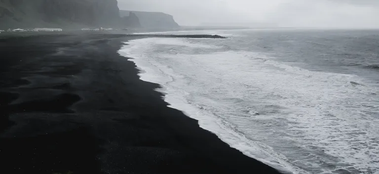 Plage de sable noir bordée de falaises, vagues blanches sous un ciel couvert