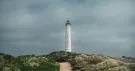 Phare blanc au bout d’un sentier de sable entre les herbes des dunes, sous un ciel gris