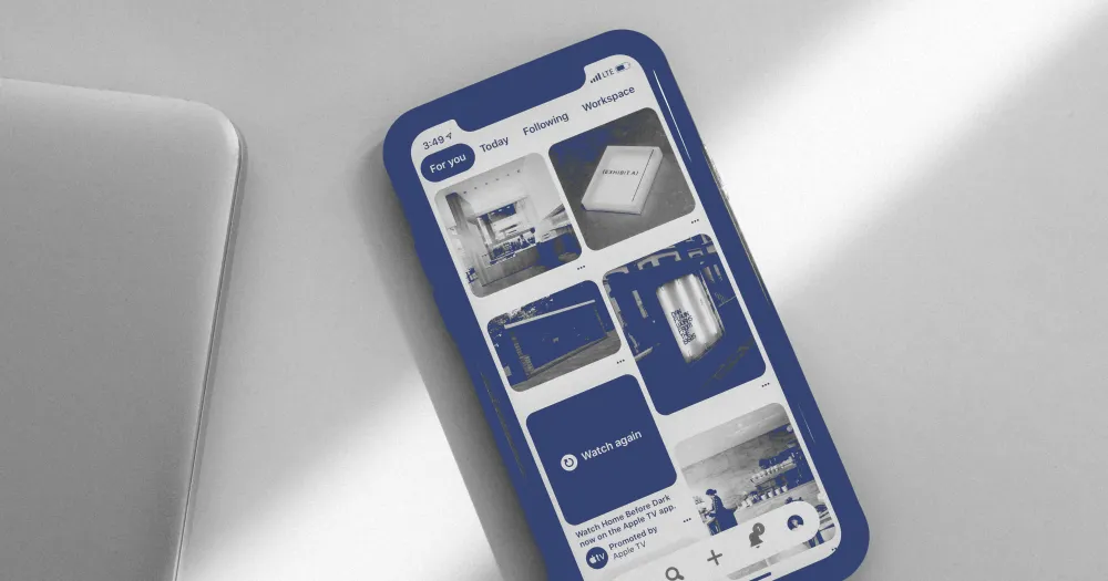 Smartphone showing an app with blue thumbnails, beside a laptop on a light grey desk