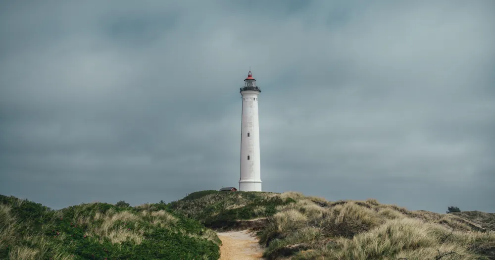 Phare blanc au bout d’un sentier de sable entre les herbes des dunes, sous un ciel gris